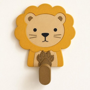 May include: A wall-mounted hook shaped like a lion's head with a golden yellow mane, a light tan face, and black facial features. A brown paw is positioned at the center, extending downward into a vertical, oval-shaped hook for hanging items.