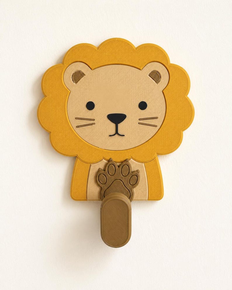 May include: A wall-mounted hook shaped like a lion's head with a golden yellow mane, a light tan face, and black facial features. A brown paw is positioned at the center, extending downward into a vertical, oval-shaped hook for hanging items.