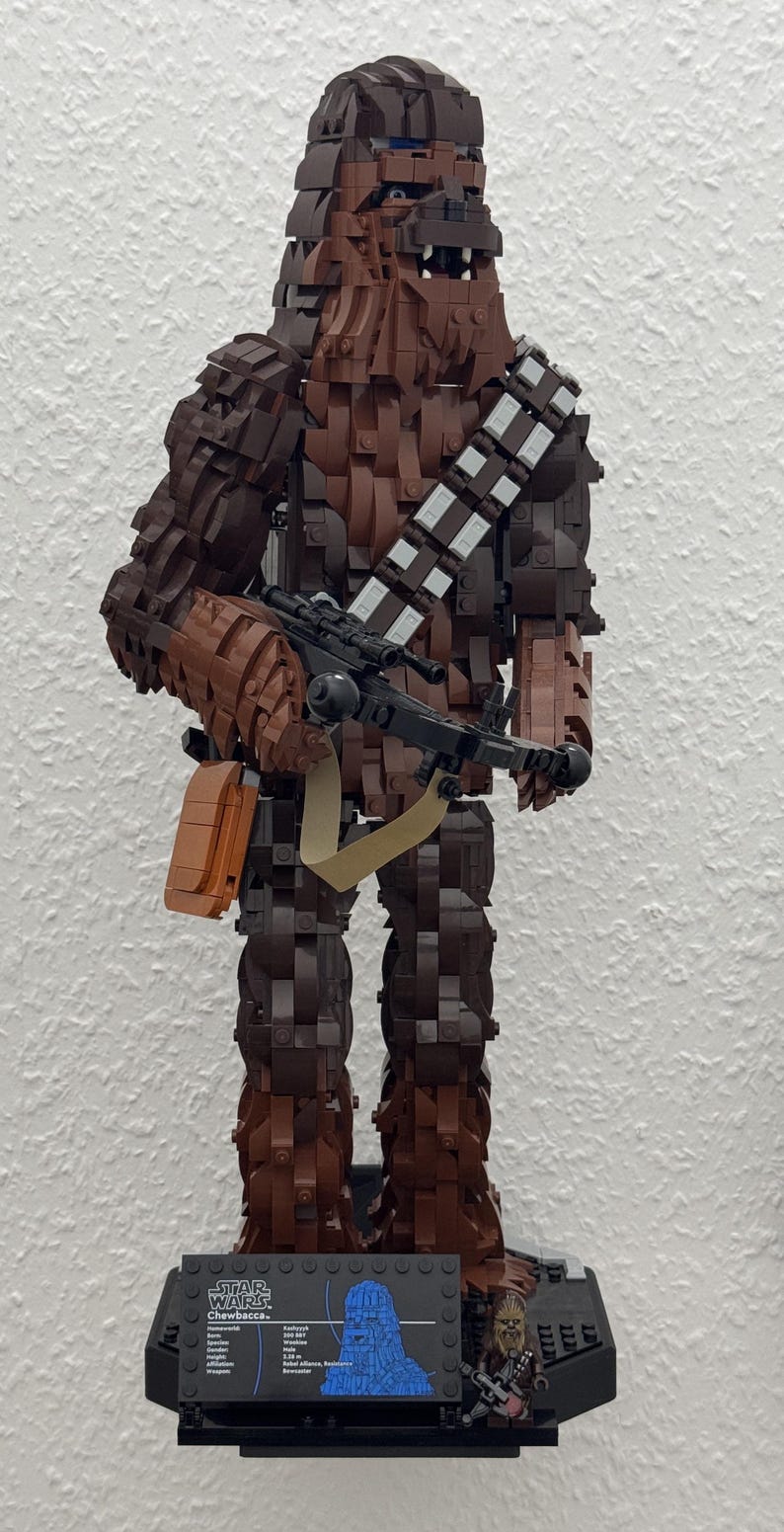 May include: A Lego sculpture of Chewbacca from Star Wars, constructed from various shades of brown Lego bricks. The figure holds a black weapon and has a bandolier. The base includes a character profile and the Star Wars logo.