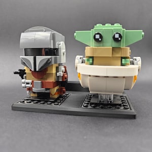 Może przedstawiać: Lego minifigurka Mandalorianina stoi obok Lego minifigurki Grogu, znanego r&oacute;wnież jako Baby Yoda, w statku kosmicznym. Obie minifigurki stoją na czarnej podstawie.