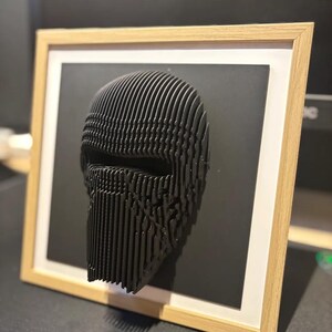 Puede incluir: Una escultura en 3D impresa en negro de una cara, enmarcada en un marco de madera clara. La escultura est&aacute; hecha de muchas capas de material delgado, creando un efecto texturizado.