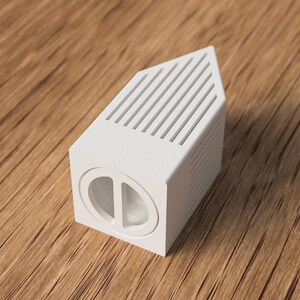May include: A white, geometric object with a rectangular base and a slanted top featuring parallel lines. The front of the object has a circular design with a vertical divider. The object is set on a wooden surface.