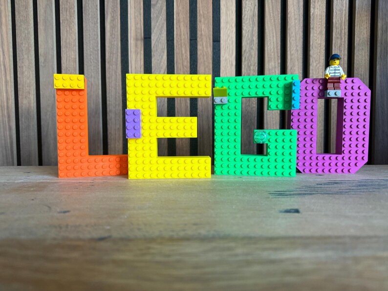 May include: The word "LEGO" is spelled out in large, colorful building blocks. The letters are orange, yellow, green, and purple. A small LEGO figure stands on top of the letter "D". The background is a wooden wall.