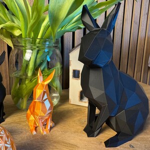 May include: Three geometric rabbit sculptures, one black, one orange, and one white, sitting on a wooden surface. The black rabbit is the largest and is in the foreground. The orange rabbit is in the middle and the white rabbit is in the background.