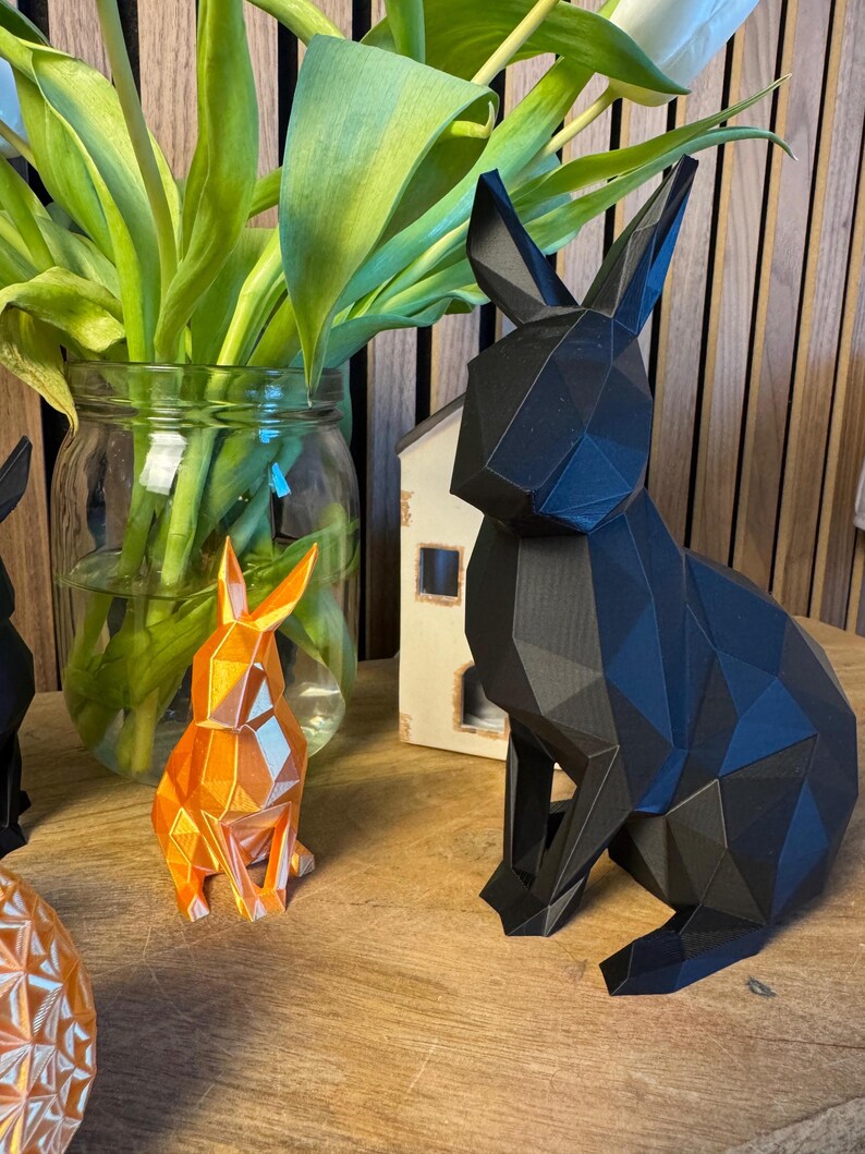 May include: Three geometric rabbit sculptures, one black, one orange, and one white, sitting on a wooden surface. The black rabbit is the largest and is in the foreground. The orange rabbit is in the middle and the white rabbit is in the background.