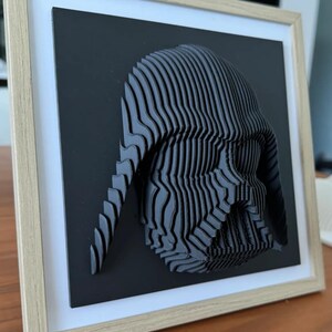 Puede incluir: Una pieza de arte de pared impresa en 3D en blanco y negro de la cabeza de Darth Vader en un marco de madera.