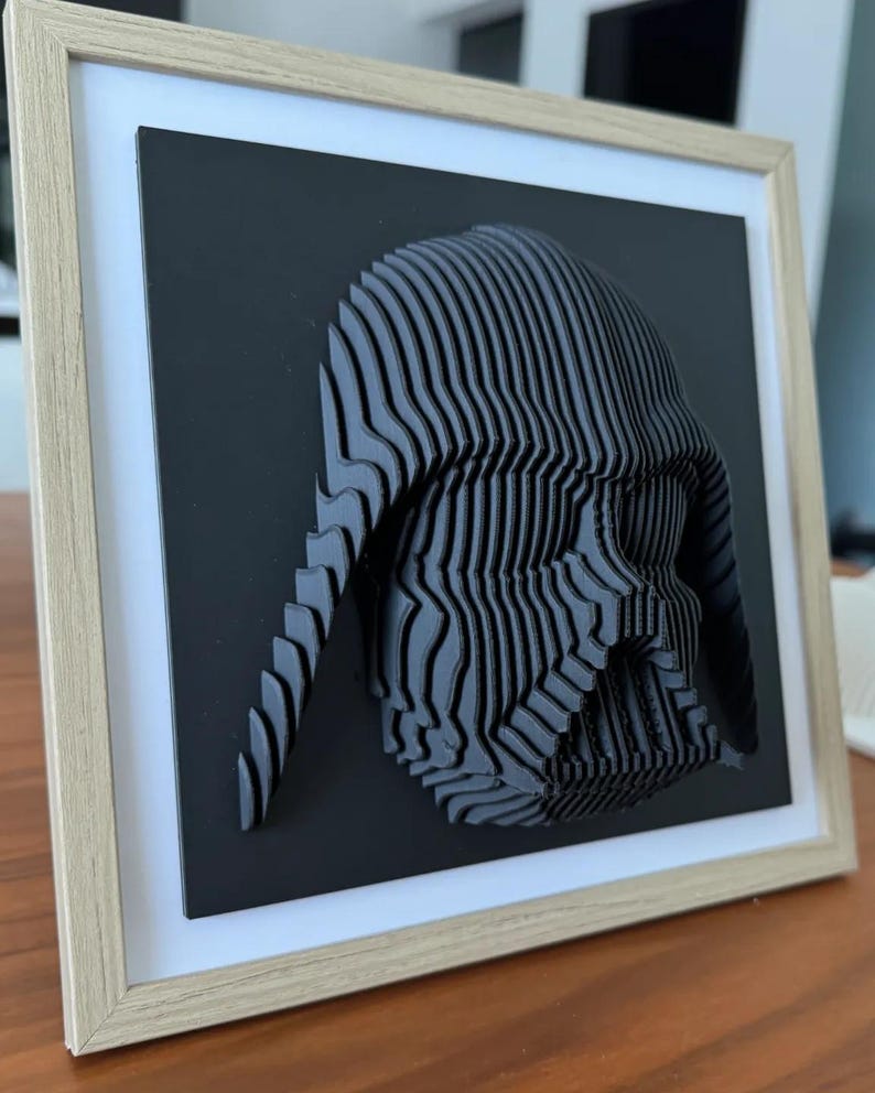 Puede incluir: Una pieza de arte de pared impresa en 3D en blanco y negro de la cabeza de Darth Vader en un marco de madera.