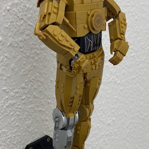 May include: A gold-colored Lego C-3PO figure from Star Wars, mounted on a black base. The figure has a silver leg and is accompanied by a small C-3PO figure and a Star Wars information plaque.