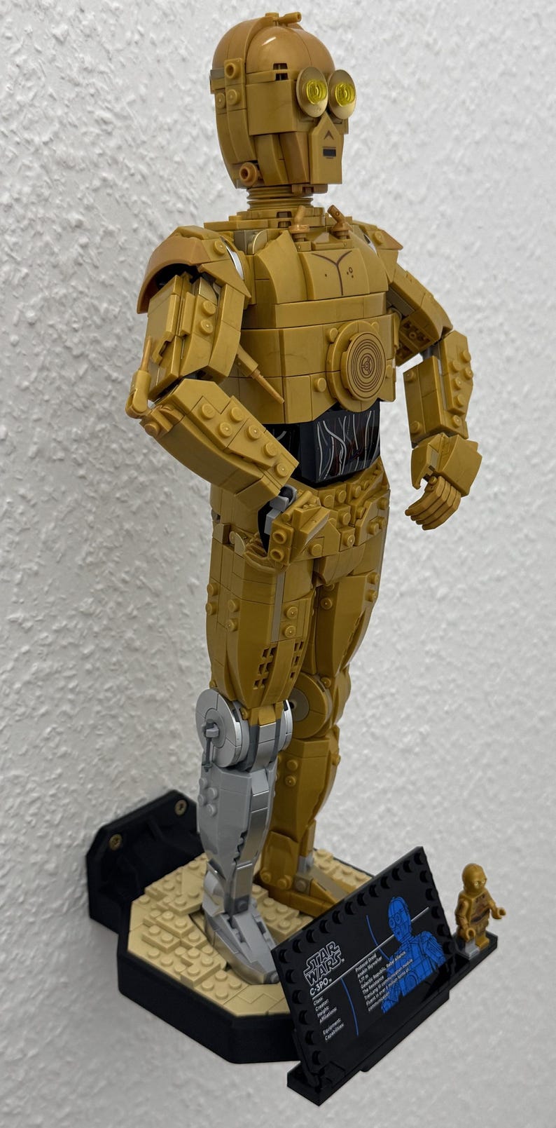 May include: A gold-colored Lego C-3PO figure from Star Wars, mounted on a black base. The figure has a silver leg and is accompanied by a small C-3PO figure and a Star Wars information plaque.