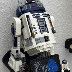 May include: A detailed LEGO model of the Star Wars character R2-D2, featuring white, blue, and gray components. The model is mounted on a black base with a display plaque. A smaller R2-D2 model is also present.