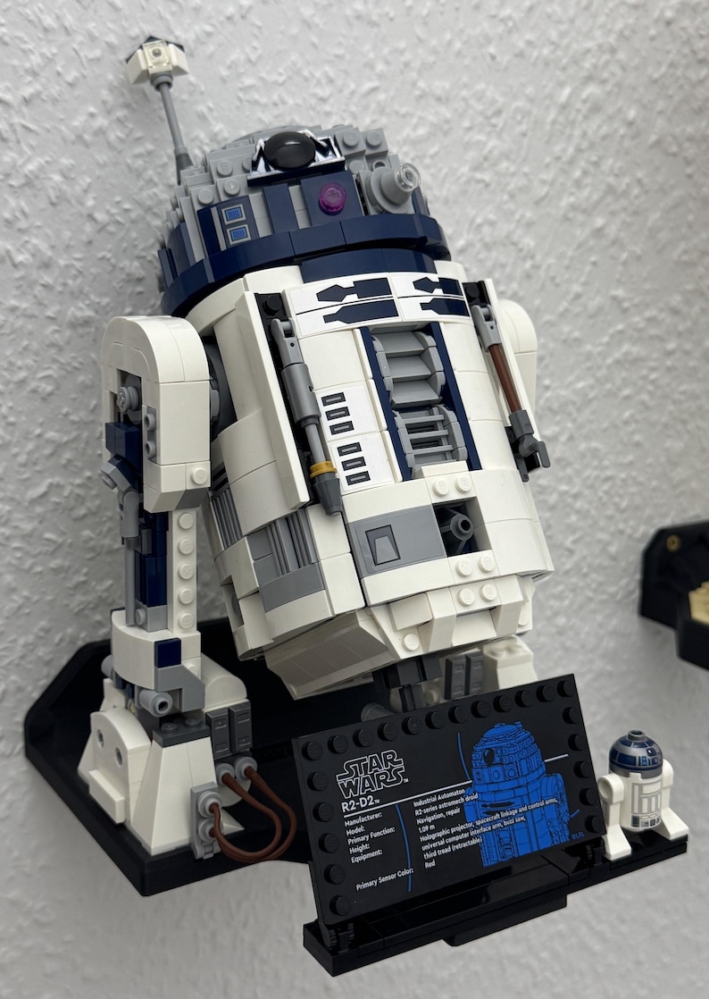 May include: A detailed LEGO model of the Star Wars character R2-D2, featuring white, blue, and gray components. The model is mounted on a black base with a display plaque. A smaller R2-D2 model is also present.