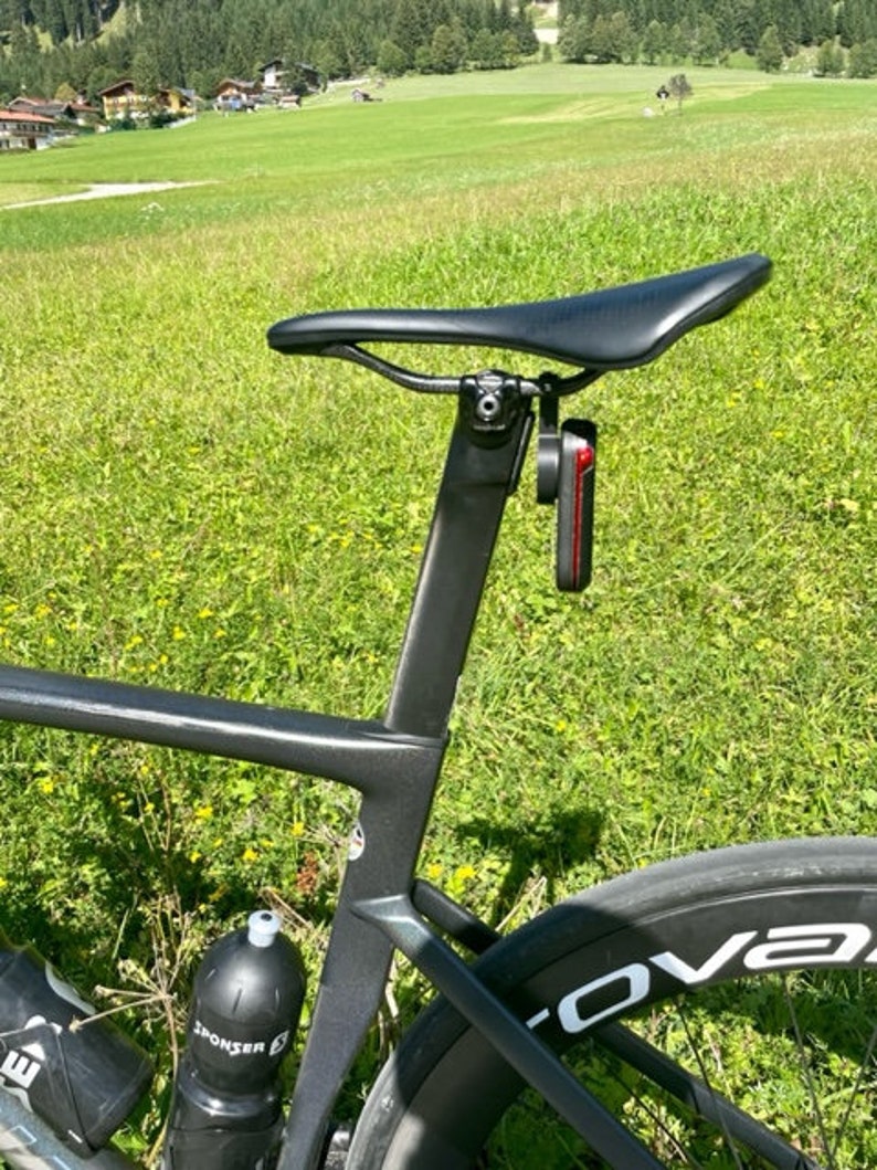 Może przedstawiać: Zbliżenie czarnego siodełka i ramy roweru z przymocowanym czerwonym światłem. Rower jest ustawiony na tle zielonej trawy i odległego pasma g&oacute;rskiego. Widoczna jest butelka na wodę.