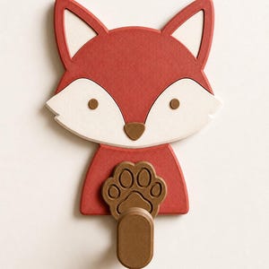 May include: A wall-mounted hook shaped like a fox head with a red and white face, brown eyes, and a brown nose. The fox has a brown paw print design on its chest, with a protruding brown cylindrical hook extending from the bottom of the paw.