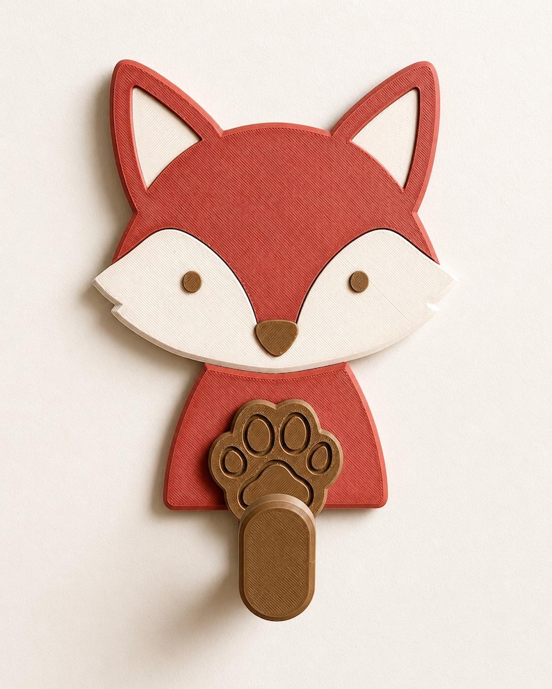 May include: A wall-mounted hook shaped like a fox head with a red and white face, brown eyes, and a brown nose. The fox has a brown paw print design on its chest, with a protruding brown cylindrical hook extending from the bottom of the paw.