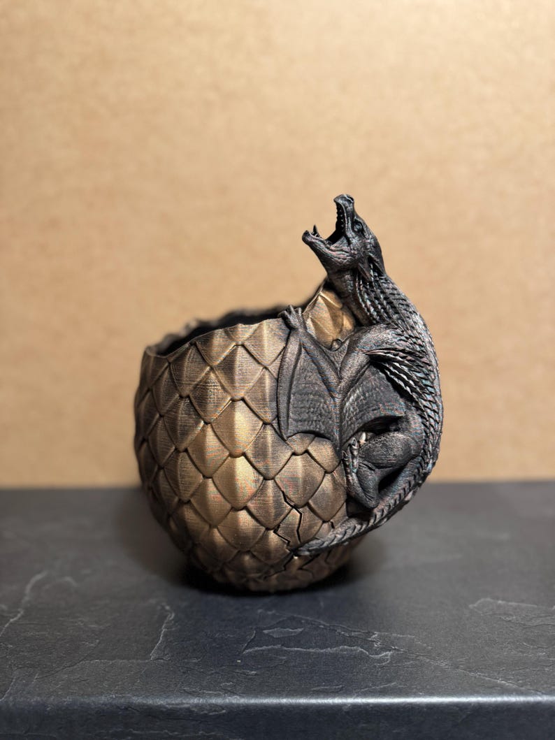 May include: A bronze-colored dragon egg planter with a detailed, textured surface. A dark gray dragon is emerging from the egg, with its mouth open. The planter is on a dark gray surface.