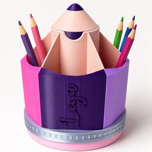 May include: A multi-colored pencil-shaped desk organizer featuring a central compartment and outer slots for stationery. The exterior is segmented in pink, dark purple, and light purple, with the name Leya embossed in script on the dark purple section.