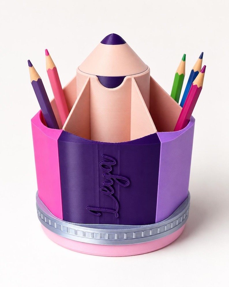 May include: A multi-colored pencil-shaped desk organizer featuring a central compartment and outer slots for stationery. The exterior is segmented in pink, dark purple, and light purple, with the name Leya embossed in script on the dark purple section.