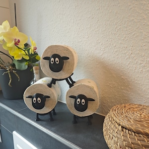 May include: Three black sheep toilet paper holders with white toilet paper rolls. The holders are stacked on top of each other. The sheep have black eyes and a black nose. There is a wicker basket in the background.