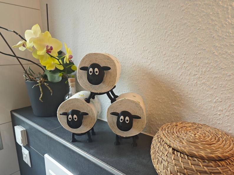 May include: Three black sheep toilet paper holders with white toilet paper rolls. The holders are stacked on top of each other. The sheep have black eyes and a black nose. There is a wicker basket in the background.