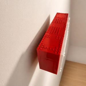 May include: A red and white rectangular object constructed from interlocking plastic bricks, mounted on a white wall. The red section is textured with studs, while the white side is smooth. The object casts a shadow.