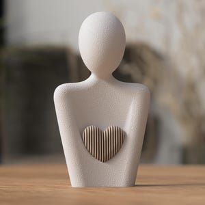 May include: A white, minimalist sculpture of a human figure with a heart-shaped cutout. The heart is made of vertical wooden slats. The sculpture has a textured surface and is set on a wooden table.