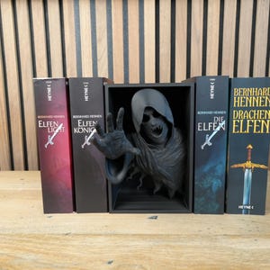 May include: A display of fantasy books with a dark, gothic theme. The books, with titles like "Elfen Licht" and "Drachen Elfen," are arranged with a dark sculpture of a skeletal figure reaching out. The books are in English and German.