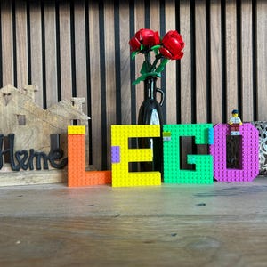 May include: A wooden house-shaped decoration with the word "Home" in black script. The word "LEGO" is spelled out in colorful blocks: orange, yellow, green, and purple. A small LEGO figure sits atop the letter "O". A black vase holds red roses.