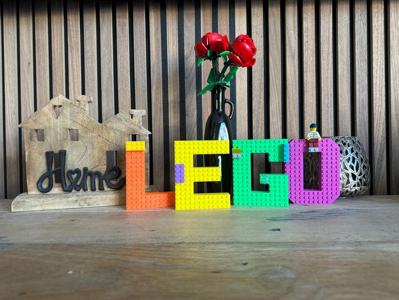 May include: A wooden house-shaped decoration with the word "Home" in black script. The word "LEGO" is spelled out in colorful blocks: orange, yellow, green, and purple. A small LEGO figure sits atop the letter "O". A black vase holds red roses.