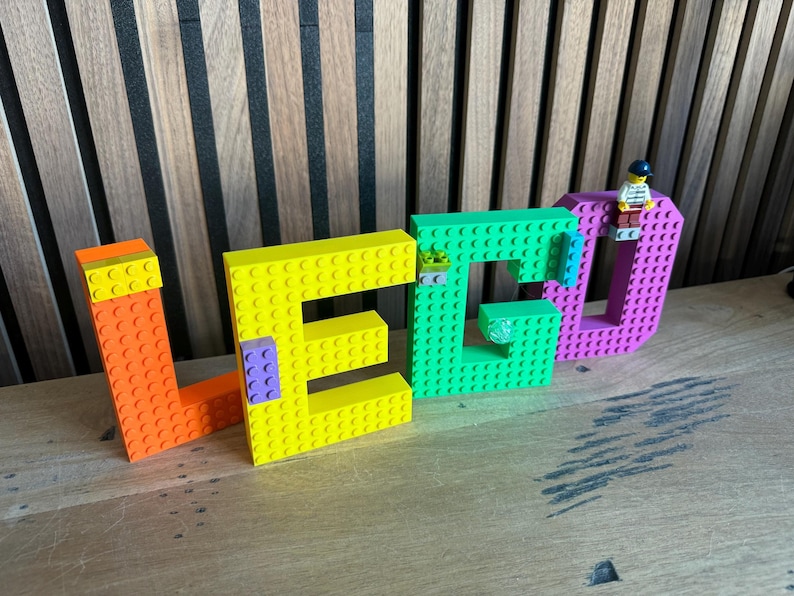 May include: The word "LEGO" spelled out in large, colorful building blocks. The letters are orange, yellow, green, and purple. A small LEGO figure stands atop the letter "D". The blocks are on a wooden surface.