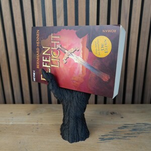 May include: A black, hand-shaped book holder supporting a book titled "Elfen Licht" by Bernhard Hennen. The book cover features a sword illustration against a red and orange background. The holder is made of a dark, textured material.