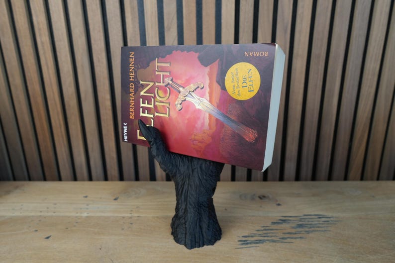 May include: A black, hand-shaped book holder supporting a book titled "Elfen Licht" by Bernhard Hennen. The book cover features a sword illustration against a red and orange background. The holder is made of a dark, textured material.
