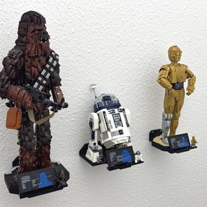 May include: Three Star Wars LEGO figures: Chewbacca, R2-D2, and C-3PO. Chewbacca is brown and holding a blaster. R2-D2 is white and blue. C-3PO is gold. Each figure is mounted on a black base with a graphic.