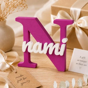 May include: A magenta-colored capital letter N with the white lowercase name nami attached to the front. The letter stands upright and features a smooth, matte finish. The name is written in a rounded, sans-serif font across the center of the letter.