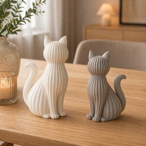 May include: Two ceramic cat figurines. One is white, the other is gray. Both are sitting upright with their tails curled. The cats have vertical ribbed detailing and pointed ears. They are placed on a light brown wooden table.