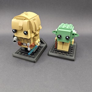 Może przedstawiać: Dwie minifigurki Lego, jedna przedstawiająca Luke'a Skywalkera, a druga Yodę, są wystawione na czarnych podstawkach. Luke trzyma niebieski miecz świetlny. Yoda nosi brązową szatę.