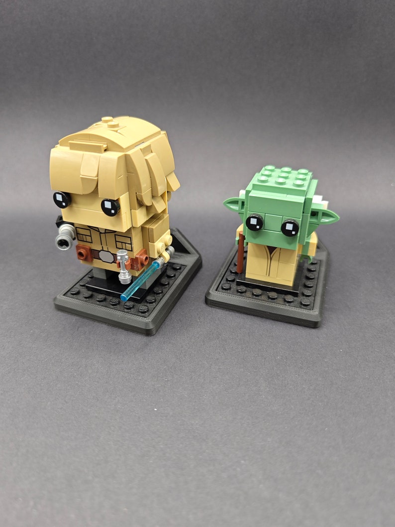 Może przedstawiać: Dwie minifigurki Lego, jedna przedstawiająca Luke'a Skywalkera, a druga Yodę, są wystawione na czarnych podstawkach. Luke trzyma niebieski miecz świetlny. Yoda nosi brązową szatę.