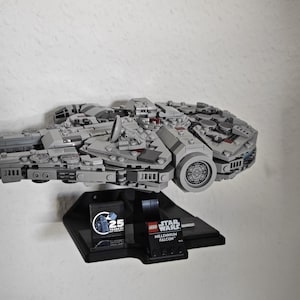 Könnte beinhalten: Ein graues LEGO Millennium Falcon Raumschiff auf einem schwarzen Displayständer. Der Ständer trägt das LEGO Star Wars 25-Jahre-Jubiläumslogo und die Worte "Millennium Falcon".