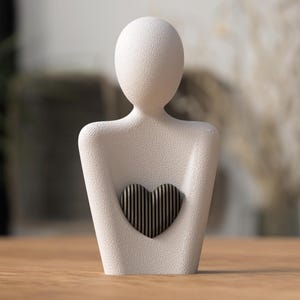 May include: A white, textured figurine in the shape of a person, holding a heart-shaped object. The heart is made of vertical, dark-colored lines. The figurine is on a wooden surface, with a blurred background.