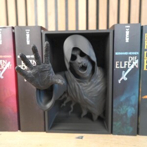 May include: A dark grey sculpture of a hooded figure with an outstretched hand, emerging from a black box, flanked by book spines. The books have titles in German, with a sword and a cross design.