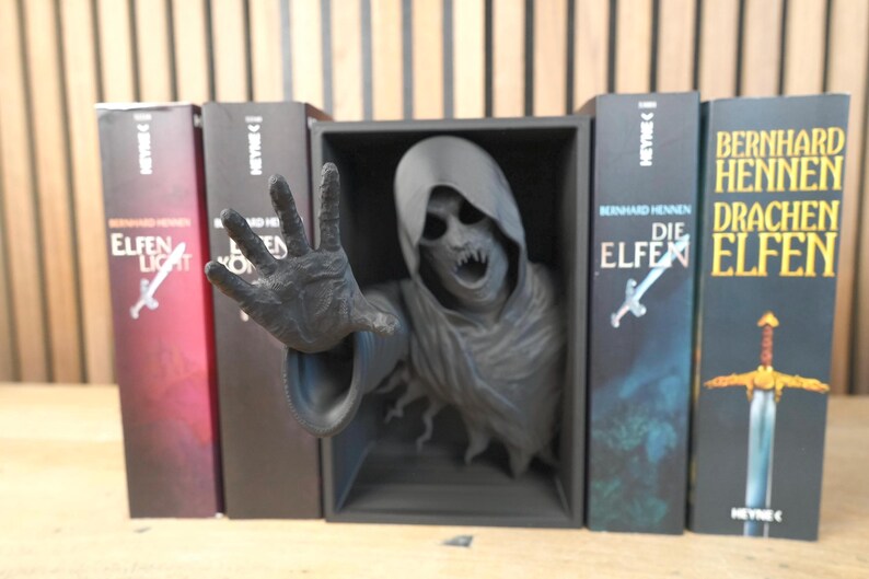 May include: A dark grey sculpture of a hooded figure with an outstretched hand, emerging from a black box, flanked by book spines. The books have titles in German, with a sword and a cross design.