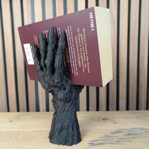 May include: A black, stylized book holder shaped like a gnarled hand, supporting a book with a burgundy cover. The book's title is partially visible, with the word "HEYNE" at the top. The holder is on a wooden surface.