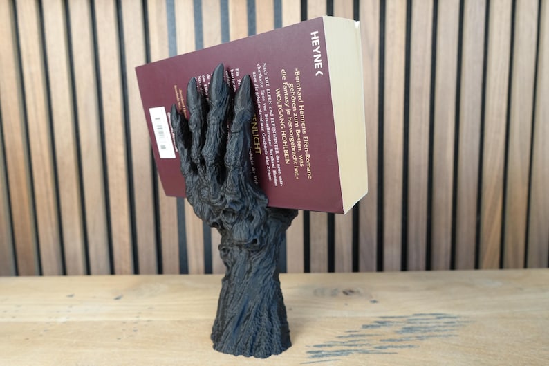 May include: A black, stylized book holder shaped like a gnarled hand, supporting a book with a burgundy cover. The book's title is partially visible, with the word "HEYNE" at the top. The holder is on a wooden surface.