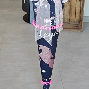 Puede incluir: Un cono escolar azul y blanco con un dise&ntilde;o de unicornio y pompones rosas. El cono tiene una base blanca con el nombre "Leya" escrito en letras rosas. El cono est&aacute; decorado con tul rosa y una cinta rosa.