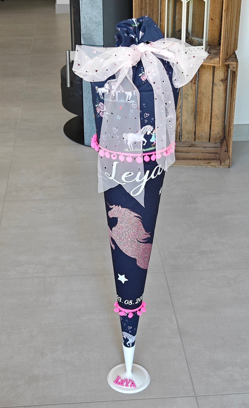 Puede incluir: Un cono escolar azul y blanco con un dise&ntilde;o de unicornio y pompones rosas. El cono tiene una base blanca con el nombre "Leya" escrito en letras rosas. El cono est&aacute; decorado con tul rosa y una cinta rosa.