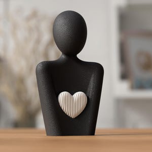 May include: A black figurine of a person holding a heart. The heart is white with vertical lines. The figurine has a textured surface and is displayed on a wooden table.