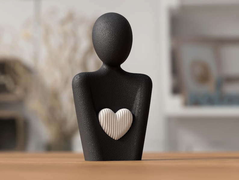 May include: A black figurine of a person holding a heart. The heart is white with vertical lines. The figurine has a textured surface and is displayed on a wooden table.