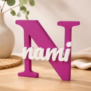 May include: A large, magenta-colored capital letter N made of a solid material. The white, lowercase name "nami" is attached horizontally across the center of the letter N. The object stands upright on a flat surface, displaying a smooth finish.