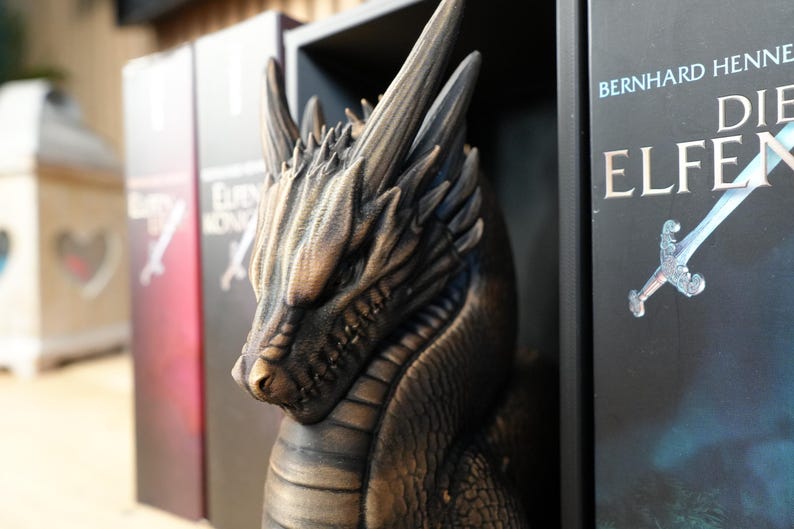 May include: A detailed bronze-colored dragon sculpture with textured scales and sharp horns. The dragon is positioned in front of a bookshelf with book titles visible. The books have a fantasy theme.
