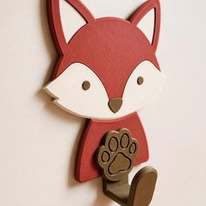 May include: A wall-mounted hook shaped like a fox head with a red face, white cheeks, and brown eyes and nose. The fox is holding a brown paw print emblem, and a brown hook extends from the bottom, providing a functional storage solution.