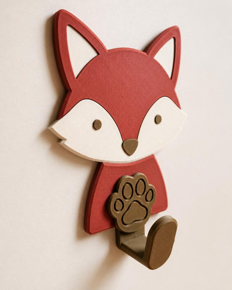 May include: A wall-mounted hook shaped like a fox head with a red face, white cheeks, and brown eyes and nose. The fox is holding a brown paw print emblem, and a brown hook extends from the bottom, providing a functional storage solution.